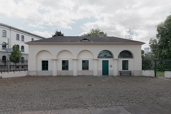 View of the front of a cream white house with five arches in a row around four windows and a door in the arch second from the right. It is a one-floor house with a bonnet roof covered in grey tiles. To the right we can see a low chimney and in the center of the roof a narrow triangular window. Between the arches are four round pillars, and the lower part of the facade is covered in light grey rectangular stones up to the windows. The four windows are rectangular and divided into six square sections by emerald green frames. The door is painted in the same green. Above the door and above the window in the arch to the very right, semicircular windows make up the arches. They are divided into sliced sections outlined in the same green framing. In front of the arch to the right stands a wooden bench. In front of the house, taking up about half of the picture, is an open cobble-stoned space. To the left and right of the house leading up to the facade runs a low wall with a ornamental black metal fence. On the left side, there is gate in the wall made from black ornamental metal, which is open. It leads to a garden or green area with trees, and a light grey tower is visible in the distance. Behind the fence on the left hand of the house, we can see a white three-storage neighboring building, a tree and street signs. The sky above the house is blue with white clouds.