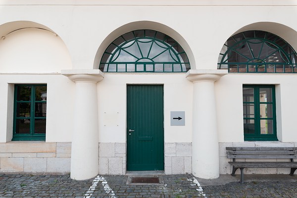 Close-up view of the door and two windows of the cream white building in the previous image. In the center of the picture we can see a green door with the handle to the left. Next to it a grey sign with a black arrow pointing to the right has been mounted on the facade. Above the door we can see a semicircular window taking up the arch, which is divided into geometrical sections in green frames emanating from the center. In the window glass, we can see the blue sky and white clouds reflected. In front of the door is a mental door grille, and on the cobble stone we can see some white lines. To the right of the door, in the next arch, we can see an identical semicircular window that reflects bits of the sky and shows some of the interior of the building. Below it, there is a rectangular window divided into six square window panes in green frames through which we can glimpse interior walls of the building and an open window. In front of the window, up against the wall, stands a wooden bench. In the arch left of the door, we can see an identical rectangular window that reflects a black metal fence. Between the arches are round, semicircular pillars.