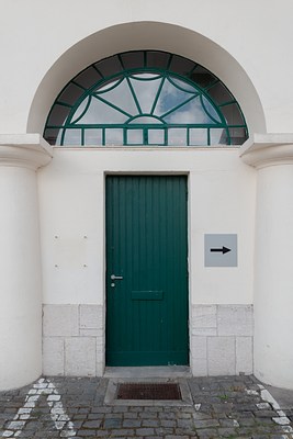 Close-up of the previous image. In the center, we can see a green door with the handle to the left. Next to it a grey sign with a black arrow pointing to the right has been mounted on the facade. Above the door we can see a semicircular window taking up the arch, which is divided into triangular and square window panes in green frames emanating from the center. In the window glass, we can see the blue sky and white clouds reflected. In front of the door is a mental door grille, and on the cobble stone we can see white lines.