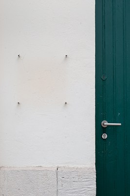 Close-up of the previous image. A cream colored plaster facade takes up two thirds of the picture from the left. To the right, we see a part of a green door with a metal door handle and key hole. On the wall we can see four sign mounts making up a square. The sign is absent. Below it, in the lower part of the picture, the facade is covered in light stones.