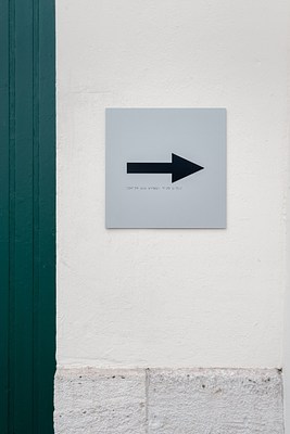 Close-up of the grey, square sign on cream plaster facade in previous images. In the center of the sign, a black arrow points to the right. Below it, there is writing in braille. To the left of the sign, we can see parts of a green door. Below the sign, in the lower part of the picture, the facade is covered in light stones.