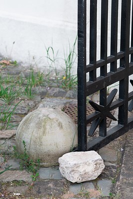 Close-up of the stones and gate in previous image. In the upper part of the picture we can see a white stone wall, crossing the picture plane diagonally. In front of it, we can see cobble-stone clad ground. Weeds break through the cobble-stone and a metal water drain is visible. To our right, we can see the lower left corner of a black metal door, entering the picture frame diagonally from the right. It consists mostly in vertical beams with a few horizontals. At the bottom, we can see parts of a star-shaped ornamental pattern. The door is held open by a light natural stone, possibly quartz, roughly cut in a cubic shape. Just behind it, we can see a larger dome-shaped stone made from casted concrete. It seems to be functioning as a door stopper, and the gate sits wedged between the two stones.. . . 