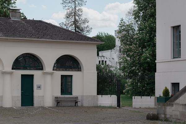 View of the cream white building with arches and tiled bonnet roof in previous images but from an angle. We can see the green door, and to its right, the window with the wooden bench. To the right in the picture, we can see parts of a taller white building and a staircase in stone leading up to its entrance to the right, out of frame. On top of the staircase stands a plant, and on the cobble stone below, lies a large rounded stone to the left. On the tall white building, we can see two rectangular windows in green frames, one on the ground floor and the other above it on the first floor. Between the two houses run a low white wall in stone with a black metal fence in ornamental style. In its middle, we can see an open gate leading into a garden with green grass and trees with white flowers. In the distance, behind the foliage, we can see parts or a large building light grey with a tower. Behind the lower building to the left, we can see the tops of two trees. The sky is blue with white clouds.