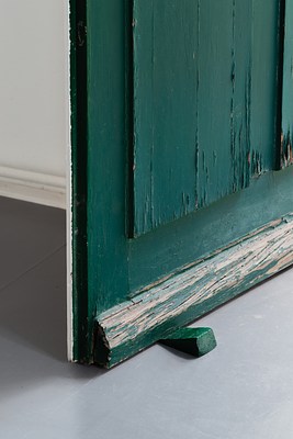 Close-up view of the lower part of the green door in previous images, swung open into a room with grey floor and white walls. The door takes up about two thirds of the picture. Its vertical edge crosses the picture from the top to about two thirds down, while its horizontal edge near the floor disappears out of the picture frame, diagonally to the right. The door is made from wood and has been painted in an emerald green. The paint is chipping, and near the floor we can see the wood exposed. In the background, we can see parts of a white wall and base moulding. The floor is grey, and as the light seems to be coming from the door opening to the right out of frame. The door is casting shadows to its left. On the vertical edge of the door we can see a white stripe, suggesting the inside of the door might be painted in white. The door is held open by a small wooden wedge painted in a similar green.