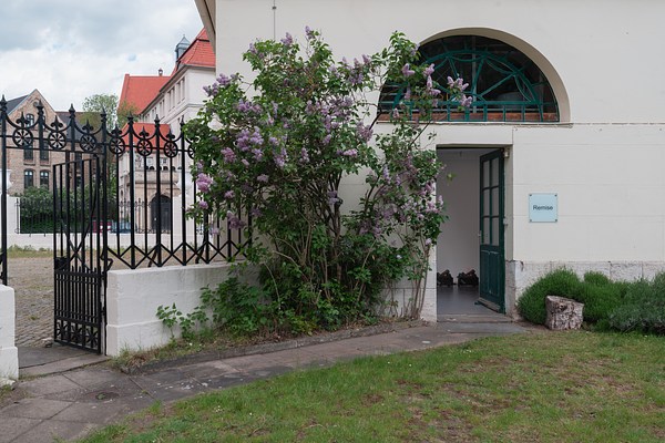 The same ornamental fence and gate alongside creme colored house as in previous images, but photographed from a different angle. In the left half of the image, we can see the low, white stone wall with the black metal fence and gate entering the frame from the lower left, extending diagonally in the picture to the middle where it meets the cream colored building. The fence and gate consists mostly in vertical beams, with some running horizontally. At the bottom of the fence, we can see a zig-zag pattern and at the top, a pattern of circles with crosses inside, and the top of the fence is made out from spear-shaped pinnacles. The gate has a similar design but ends in a vertical beam. At its bottom we can see a pattern of star-shaped ornaments. From the gate, a stone-paved path turns sharply to the left and leads up to a doorway in the cream colored house. The door of the house is open. We can see a grey floor and what appears as sculptures made from burning logs. To the left of the door, we can see a tree with green leaves and purple-pink flowers in bloom. To the right of the door, on the wall, we can see a light grey square sign with the text "Remise." In front of it, on the ground, stands a tree stump. Above the door we can see an arched window with green frames.