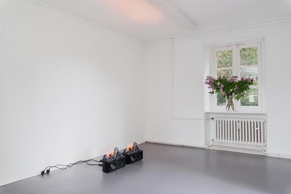 Installation view of a room with white walls and ceiling and a grey floor. The two walls form a corner at the center of the picture. The left wall is empty whereas the right one has a window at its center with a heater below. In the window stands a transparent glass vase with water and a bouquet of branches with green leaves and purple flowers cut from the tree in the garden outside. The window is rectangular and made up from six square window panes. Its frame is painted white. The bouquet takes up about half of its height and entire width. Through the window we can see green foliage. On the floor in front of the left wall sit two identical small black boxes plugged into electrical sockets on the wall to their left. On their top, identical configurations of logs appear burning in flames. Through small slits in the boxes, wires and electrical components are visible. In the white ceiling directly above, we can see a warm light reflected.