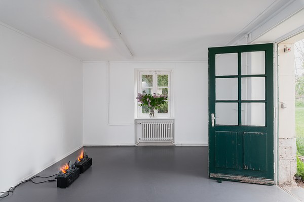 Installation view of the same room as in previous two images with grey floor, three white walls, white ceiling, window and green door. We are facing a wall with the window. Directly to our left is an empty wall, and to out right, a wall with a green door swung open. On the floor in front of the left wall sit two identical small black boxes plugged into electrical sockets on the wall to their left. On their top, identical configurations of logs appear burning in flames. Through small slits in the boxes, wires and electrical components are visible. In the white ceiling directly above, we can see a warm light reflected. The window on the center wall is rectangular and made up from six square window panes. Its frame and ledge is painted white. Directly beneath it, we can see a white radiator. On the window ledge stands a transparent glass vase with water and a bouquet of branches with green leaves and purple flowers cut from the tree in the garden outside. Through the window we can see green foliage. The green door to the right in the image stands wide open. It is made from wood and has been painted in an emerald green. The paint is chipping, and near the floor we can see the wood exposed. In the upper half, it has a rectangular window in the same design as the window in the center wall, made up from six square window panes. The door handle is to the left, and made from metal. The door is held open by a small wooden wedge painted in a similar green. Through the doorway, we can see the thickness of the exterior wall of the building, and a sliver of green grass, foliage, and light sky.