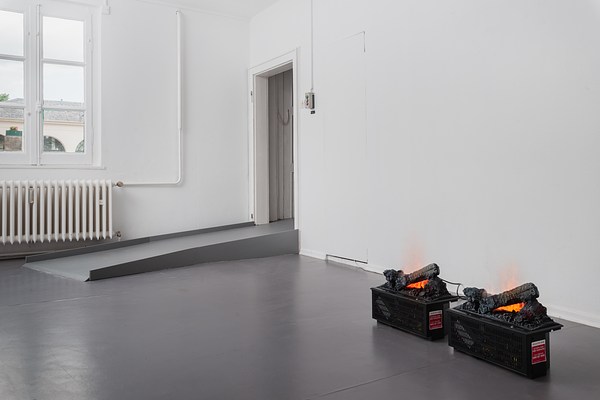 Installation view of the same room with white walls and grey floor as in previous images but from a different angle. From this viewpoint, we can see two walls meeting in a corner at the center of the image where a doorway is located on the right wall. From the doorway, a ramp in the same color as the floor juts out straight from the doorway, pointing to the left in the image. Through the doorway we can glimpse parts of a closed white door. Next to the door, on the wall, we can see a small grey, rectangular box, with a green, grey, and red button vertically arranged. On the left wall, a window is located above the grey floor ramp. The window is rectangular and made up from six square window panes. Its frames are painted in white. Directly beneath it, we can see a white radiator, and a L-shaped pipe leading into it from the ceiling. Through the window we can see a light grey sky, and the upper part of a cream colored building with arched windows and a grey, tiled roof. The light from the window is reflected in the polished, grey floor. In the right bottom corner of the image, in front of the right wall, two identical small black boxes sit directly on the floor, plugged into electrical sockets on the wall. On their top, identical configurations of logs appear burning in flames. Through small slits in the boxes, wires and electrical components are visible.