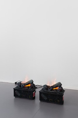 Close-up of the two identical small black boxes as in previous images, sitting directly on the floor, plugged into electrical sockets on the wall. On their top, identical configurations of logs appear burning in flames. Through small slits in the boxes, wires and electrical components are visible.