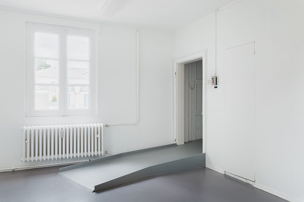 Installation view of the same room with white walls and grey floor as in previous images. We can see two walls meeting in a corner at the center of the image where a doorway is located on the right wall. From the doorway, a ramp in the same color as the floor juts out straight from the doorway, pointing to the left in the image. Through the doorway we can glimpse parts of a closed white door. Next to the door, on the wall, we can see a small grey, rectangular box, with a green, grey, and red button vertically arranged. On the left wall, a window is located above the grey floor ramp. The window is rectangular and made up from six square window panes. Its frames are painted in white. Directly beneath it, we can see a white radiator, and a L-shaped pipe leading into it from the ceiling. Through the window we can discern a light grey sky, and the upper part of a creme colored building with arched windows and a grey, tiled roof. The light is very bright, and the view through the window appear over exposed. The white light from the window is reflected in the polished, grey floor and on the ramp.