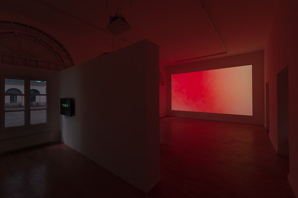 Installation view of a large dark room with two walls that meet in the a corner slightly to the right of the center of the image. At the beginning of the left wall, we can see a rectangular window divided into six square window panes. The window glass appears to be covered in a foil that prevents light from entering the room. Through the dark foil, we can still discern a building with arches and windows on the other side of a cobble-stone courtyard. Above the rectangular window, we can glimpse a semicircular window of which the window panes have either been painted or covered in opaque foil. No light enters. To the right of the window, a temporary exhibition wall juts out perpendicular to the wall. On it, close to the window, we can see a black screen mounted at eye-height. On the screen we can read "Strobe On" in bright green capital letter text. To the right of the wall, we can see the rest of the room. A large projection takes up the entire wall opposite the temporary exhibition wall. The projection is a color field fading from a warm pinkish red, to orange, and pale yellow. The wooden floor in front of the projection and and the white walls and ceiling around it reflects the warm light.