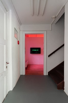 Installation view of a hallway with white walls, white ceiling, and a dark grey floor, leading into a room with red light. Directly to our left we can see a closed, white door with metal handle. On the left wall, further down the hallway, we can see a red fire alarm box and what looks like a fire escape plan mounted on the wall. From the box, a cable runs across the wall at the end of the hallway, over to the other side, and up the ceiling. On the opposite of the fire escape plan, we can see a doorway with the start of a brown wooden staircase and handle leading to the floor above, out of frame, as well as a sliver of white wall directly to our right. In the ceiling we can see three white painted pipes, running in the same direction as the hallway, as well as a smoke detector to the right. At the end of the hallway, a doorway leads into the next room. There we can see a wooden floor, parts of a radiator to the left and a wall facing us. The wall is not a bearing wall and possibly temporary as it doesn't go all the way up to the ceiling. On the wall hangs a black screen which shows the text "STROBE OFF" in bright green. The light in the room is red.