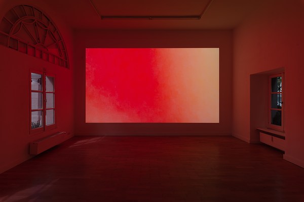 Installation view of the same a large dark room with projection as in previous image but from a different angle. On the wall directly in front of us, we can see a projection taking up the entire wall. The projection is a color field fading from a warm pinkish red, to orange, and pale yellow. In this moment, there is no text seen on the projection.