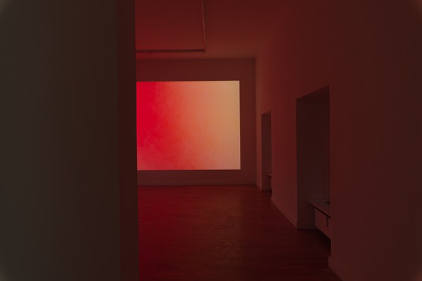 Installation view of the same large dark room with projection as in previous image but from a different angle. In the foreground to the left, a dark wall takes up about one third of the image. Behind the wall in the foreground, we can see about half of a projection that takes up the wall we are facing. The projection is a color field fading from a warm pinkish red, to orange, and pale yellow. To the right of the projection, and directly to our right, we can see a white wall with two windows. The angle does not let us see the actual windows as they are inserted into the thick walls but we can discern radiators below them. In the ceiling we can see fluorescent light tubes which are not lit. The wooden floor, and the white wall and ceiling reflects the warm red light from the projection.