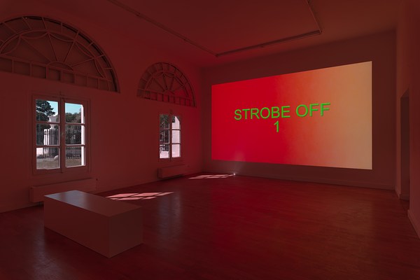 Installation view of the same a large dark room with projection as in previous image but from a different angle. We can see two walls meeting in a corner at the center of the image. On the left wall, we can see two rectangular windows made out of six square window panes each. Above them, large semicircular windows have been blacked out. Beneath them hangs white, rectangular radiators. Through the windows we can glimpse blue sky, trees and in the foreground a low white stone wall with a high ornamented metal fence in black. On the wooden floor in front of the windows, we can see the light from outside reflected in bright square patterns. On the right wall, we can see a large projection. The projection is a color field fading from a warm pinkish red, to orange, and pale yellow with the text "Strobe Off 1" in centered, bright green capital letters. In front of the projection, far down the opposite side of the room, and in the bottom left corner of the image, we can see a simple rectangular bench in white, reflecting some of the light from the windows. The wooden floor in front of the projection and and the white walls and ceiling around it reflects its warm red light.