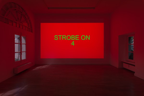 Installation view of the same a large dark room with projection as in previous image. On the wall directly in front of us, we can see a projection taking up the entire wall. The projection is a color field fading from a warm pinkish red, to orange, and pale yellow, with the text "Strobe on 4" in centered, in bright green capital letters. The wooden floor in front of the projection and and the white walls and ceiling around it reflects its warm red light. We can also see some daylight reflected in the floor coming from the window in the left wall.
