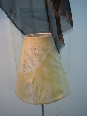 A detail view of the smaller floor lamp seen in the last few images. We can see now that paper with text has been wrapped around the lampshade, and long strips of yellowing tape have been applied over it. Sheer black fabric drapes over it from the larger shade above, which cannot be seen in this image. None of the text is readable even after zooming.