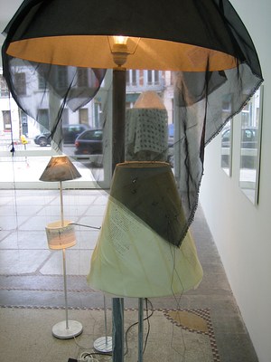 A tighter view of the two modified floor lamps. Sheer black fabric with frayed ends has been tossed over the top of one of the lampshades and drapes down over a smaller lamp next to it. Text has been stamped or printed onto the smaller lampshade. It's applied at an angle and can't be read even after zooming into the image. Other lamps and windows that look out to the street beyond the gallery can be seen through the sheer black fabric.