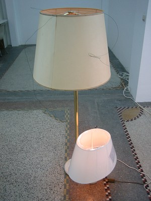An image of two other lamps among the large group of lamps in the gallery. One larger lamp has a gold stand and a tan lampshade, while a smaller lamp has a white lampshade.
