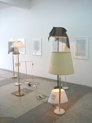 A brighter installation view of the gallery with natural light coming from our left. Along the gallery wall four framed texts hang. Floor lamps and desk lamps sit on the gallery floor in a crowd of different styles and heights. Many of their shades are draped with scarves and some of the lamp shades have texts attached. The lamps are on but can't compete with the natural light filling the space.