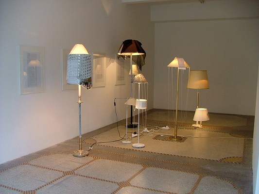 Another installation view of a gallery with white walls and a polished concrete floor in a pattern of light and middle grey, black and yellow. Along the gallery wall four framed texts hang. Floor lamps and desk lamps sit on the gallery floor in a crowd of different styles and heights. Many of their shades are draped with scarves and some of the lamp shades have texts attached. The lamps are on and throw a warm light on the gallery.
