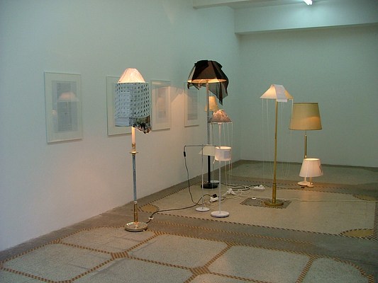 Installation view of a gallery with white walls and a polished concrete floor in a pattern of light and middle grey, black and yellow. Along the gallery wall four framed texts hang. Floor lamps and desk lamps sit on the gallery floor in a crowd of different styles and heights. Many of their shades are draped with scarves and some of the lamp shades have texts attached.