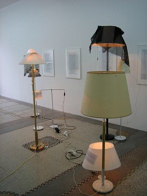 Another installation view of a gallery lit with natural light. Along the gallery wall four framed texts hang. Floor lamps and desk lamps sit on the gallery floor in a crowd of different styles and heights. Many of their shades are draped with scarves and some of the lamp shades have texts attached. The lamps are on, competing with the natural light in the gallery.