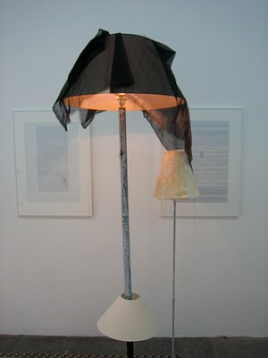 Another view of the modified lampshades taken from the other side, now facing the wall with the framed texts. The image focuses on the top half of the floorlamps. The shorter, more delicate lamp sits next to the taller lamp. Sheer black fabric drapes over the tall lampshade onto the smaller, narrower lampshade. Both stands have been painted with a layer of thin white paint that drips and swirls, not completely covering the black metal of the lamp's base.