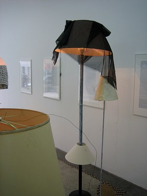 Another view of the modified lampshades taken from their other side. Over the top of another floor lamp we can see the two modified floor lamps in the last image. The shorter, more delicate lamp sits next to the taller lamp. The sheer black fabric thrown over the tall lampshade overhangs onto the smaller, narrower lampshade. Both stands have been painted with a layer of thin white paint that drips and swirls, not completely covering the black metal of the lamp's base.