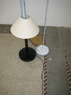 Another detail of the pair of lamps seen in the last few images. This image focuses on the bases of both lamps. On our left is a round black base with a black stem that shortly leads to a small lampshade but this shade doesn't have a bulb, it has been added like a bead to the long stem of the floorlamp. The stem of this lamp continues out of the top of the lampshade but coming out, it's been spray painted white. So the bottom of the lamp looks like a small black desk lamp with black base and tan lampshade but coming up out of the tan shade the stem continues as white up to a bulb and second shade (not seen in this detail shot.) On our right, a small round base and narrow stem have been spray painted white. The original black or gray metal shows through in places and the paint is mottled and uneven.