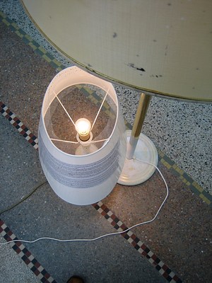 Another view of the tan and white lamps from the last image. This image is tighter, taken from above and looking down to the lightbulb in the shorter lamp. A paper with text has been wrapped around the white lampshade and secured with a piece of clear plastic. The text cannot be read even after zooming into the image. From this angle we can see that the larger floor lamp with the tan lampshade has been painted white at the base. The round metal base is white and the paint extends up the gold stand five or six inches.