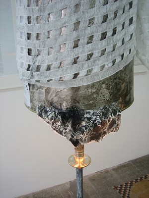 A detail of the modified floorlamp described in the last image. From this angle it is clearer that rather than two pieces of fabric attached to the bottom of the lampshade, there is a green marbled lamp shade with a fragment of marbled paper attached to one edge to make an uneven line. This has been draped with a light gray fabric woven in a loose grid. This fabric or net allows some green from the shade below to come through.