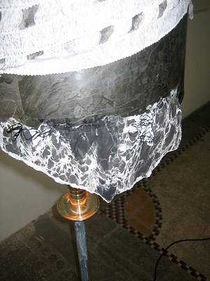 Another detail of the lampshade with a fragment of marbled paper taped to its bottom edge. Squiggles of black ink have been added in places over the pattern of white and gray marbling in the paper. The gray loose weave fabric drapes over the top of the lampshade. The image appears to be taken with a flash and all the lights stand out sharply.
