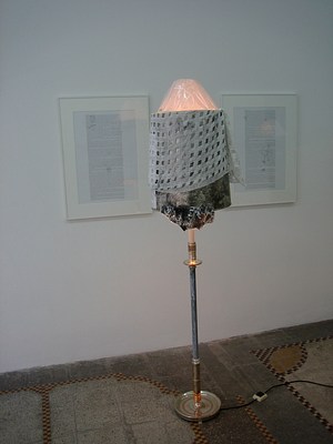 An image of a floor lamp placed between two framed text works. The lamp has a round gold base and stem and then a joint. After this joint, five or six inches up, the stem has been painted with a layer of thin white. The paint is mottled and black shows through. The lampshade at the top is white, covered by transparent plastic, as if it was fresh from a store. Around the bottom edge of the lamp two layers of fabric have been draped. The first layer has an uneven edge and is a combination of greens and black. The layer above is a light grey grid, where the weave is so wide that the fabric below shows through. The lamp is on.
