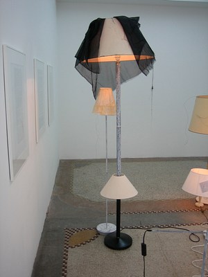 A tighter view taken from close to the gallery wall. The floor lamp in the center of the image has been modified slightly. The base of the lamp is round and black with a black pipe leading up four or five feet to a lampshade. Another, smaller lampshade has been strung along the base as if two lamps had been put together. The black base is thinly painted with white following the second shade. The white paint drips and swirls slightly, the black still partly visible. A sheer black fabric with fraying ends has been thrown over the beige lampshade at the top. Another lamp, shorter, with a narrow lampshade and a painted base, sits right next to the former floor lamp, as if in the shade of a tree. Both lamps are on.