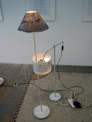 Another pairing of lamps among the group in the gallery. On our left, a tall floor lamp with a round base and stem painted white. The paint is thin and mottled. The lamp shade has been painted or stained with brown. The glow of the bulb shows through the layers of paint. A thin white string hangs in long loops from the top of the lampshade. Next to this lamp, a floor lamp with an adjustable arm is tipped to be perpendicular to its stem. The stem and cross arm are painted white. The lamp looks like it has a black metal shade, of the same material as the lamp but it has been surrounded by a traditional fabric shade. This second lamp is leaning close to the first lamp, and the stem of the first lamp is coming out through the second lampshade. The shade is a loop and the thin stem of the other floor lamp is strung through that loop so the two lamps are connected. The lampshade itself is covered with a piece of paper with text (unreadable even with zoom).