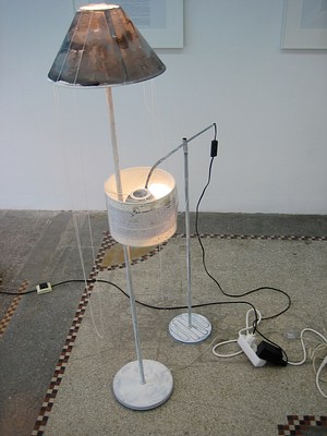 A slightly different image of two modified floor lamps. On our left, a tall floor lamp with a round base and stem is painted white. The paint is thin and mottled. The lamp shade has been painted or stained with brown. The glow of the bulb shows through the layers of paint. A thin white string hangs in long loops from the top of the lampshade. Next to this lamp, a floor lamp with an adjustable arm is tipped to be perpendicular to its stem. The stem and cross arm are painted white. The lamp looks like it has a black metal shade, of the same material as the lamp but it has been surrounded by a traditional fabric shade. This second lamp is leaning close to the first lamp and the stem of the first lamp is coming out through the second lampshade. The shade is a loop, and the thin stem of the other floor lamp is strung through that loop so the two lamps are connected. The lampshade itself is covered with a piece of paper with text (unreadable even with zoom).