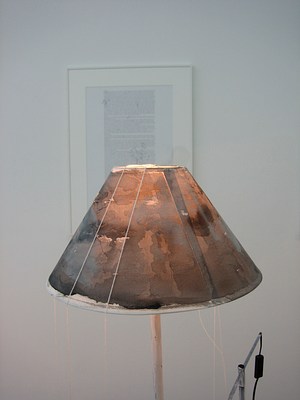 A detail of a lampshade. The lamp shade has been painted or stained in brown. The glow of the bulb shows through the layers of paint. A thin white string hangs in long loops from the top of the lampshade.