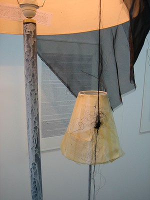 A tighter view of the two modified floor lamps. This image is taken from under the larger floor lamp so that we can see inside the lampshade. A piece of paper with text has been added to the inside, well illuminated by the glow of the lamp. The text is very small and unreadable even after zooming. There is similarly small text applied to the outside of the smaller, narrower lampshade below it. The base of the shade appears to have been spray painted with white paint that does not fully cover the original black metal stand.