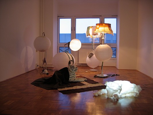 An installation view of a dim gallery space with white walls and a parquet wood floor. The back wall of the gallery is made up of windows looking out to the roof of another building. The center of the gallery is full of lamps. Four large floor lamps are clustered together in the gallery. Two of these lamps have spherical paper lamps added to their bases like beads while the their tops are illuminated with traditional lamp shapes covered with papers and tape illuminated into a patchwork surface. All four lamps have books hanging from strings down their sides and long pull cords. In the center of the image, a large stuffed tiger lies on a rug, partially covered by a blanket. To our right an inflatable lamb sculpture of translucent white is lit from within.