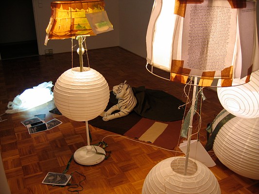 A tighter view of the floor lamps in the gallery. The image is positioned from above, close to the height of the floor lamps. From this angle we can see that pages of text have been taped to lamp shades to be easily read. Spherical paper lanterns hang from twine along the stems of the floor lamps. Photographs have been scattered along the floor next to the stuffed animal tiger lying on the rug under a blanket and the lamb sculpture/light that from this angle looks like a pile of illuminated white on the floor.