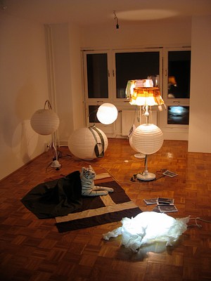 Another installation view of the gallery filled with lamps. Some lamps are turned on while others are off. The room has a cozy and dim atmosphere. The windows at the back of the gallery showing full darkness now and reflecting back the lamps in the room. The tiger sculpture on the rug and under a throw blanket appears ready to read by lamplight next to the lamb sculpture who is itself a lamp. The lamb might be a kind of half inflated creature, partially collapsed but glowing with light from within.