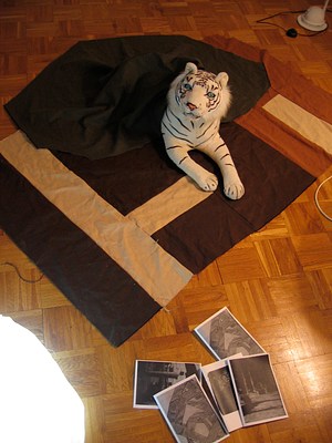 Another image taken from above. The stuffed animal tiger looks up directly at us from it's position on the carpet, covered partly with a throw blanket. Its eyes are blue. It is a white tiger with black stripes. The rug it lies on is black with strong block stripes of tan and brick red. On the floor near the rug are a pile of five pamphlets (or they appear to be pamphlets although they might be single images). To our left in the foreground, part of a white lamp brightly fills the corner of the image.