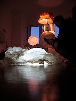 Another image taken from floor height. The gallery is much darker and only the floor lamps provide light. The lamb sculpture/lamp is a white blob in the foreground while the floor lamps provide very warm, orange light from above. On our right, a person crouches near the lamb/light while reading a pamphlet.