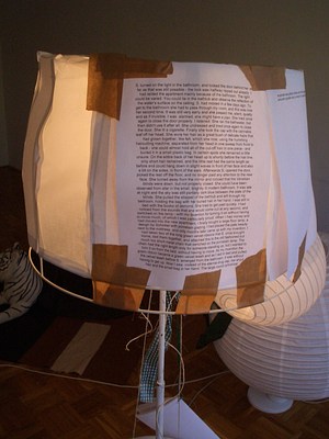 A detail of a lampshade with a text taped over it. The text reads: . "S. turned on the light in the bathroom, and locked the door behind her as far as that was still possible - the lock was halfway ripped out already. I had rented the apartment mainly because of the bathroom. The light could be varied. You could lie in the bathtub and observe the reflection of the water’s surface on the ceiling. S. dad moved in a few days ago. To get to the bathroom she had to pass through my room, and this was now her second time. It was still very early and she passed me, silent, quietly and as if invisible. I was alarmed, she might have a plan. She tried once again to close the door properly. I listened. She ran the bathwater, but then didn’t use it after all. She undressed and tried once again to close the door. She lit a cigarette. Finally she took the cap with the cannabis leaf off her head. She wore her hair as a great bush of delicate hairs that had grown together, like felt, which she now, using the humming haircutting machine, separated from her head in one sweep from front to back - she could almost hold all of the cut-off hair in one piece - and buried it in a small plastic bag. In certain spots she remained a little unsure. On the entire back of her head up to shortly before the hair line, only short hair remained, and the little rest had the same length as before and could hang down in slight waves in front of her face and also a bit on the sides, in front of the ears. Afterwards S. opened the door, picked the rest off the floor, and no longer paid any attention to the new face. She turned away from the mirror and noticed that the Venetian blinds were down, but not properly closed. She could have been observed from afar in the small, brightly lit modern bathroom. It was late at night and the sky was still partially dark blue between the slats of the bedroom, holding the bag with her buried hair in her hand. I was still in bed with the books of demons. She tried to get past quickly. I had noticed from the sounds that she would come out at any second, and switched on the lamp - with my invention for turning it on without having to move much, of which I was particularly proud. When I had money and had moved into the new apartment, I finally bought a large floor lamp, a design by Schinkel with porcelain glazing. I had placed the pale beauty next to the mattress, and only months later came up with my invention. I had taken two of the long green velvet ribbons S. once brought home, tied them together, and attached this to the old fashioned but much too short metal chain that switched on the porcelain lamp. The chain had the right length only for someone standing up, but I wanted to turn in on from the bed, without having to move. As my invention the green ribbon became a green velvet leash and as I laid in bed and pulled the velvet leash before S. emerged from the bathroom, it was without having to get up. Now I saw, instead of the eternal drug cap, her short hair and small bag in her hand. The large round cylindrical". . Another page of text is taped along the curve of the lampshade but is impossible to read.