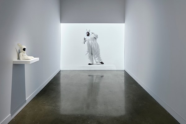 Image of a person in a bunny costume projected onto a gallery wall with a small sculpture of a bunny astronaut on a shelf on the wall to the left.&nbsp;
