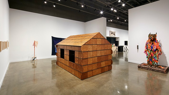Installation view featuring a wooden cabin sculpture in the foreground and a colorful bear sculpture on the left.&nbsp;