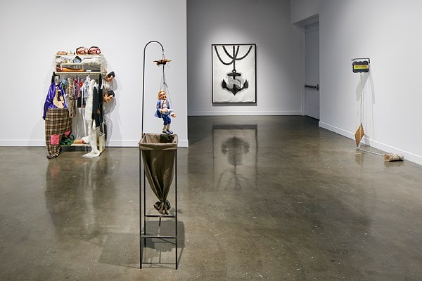 Installation view featuring a sculpture of a puppet with a laundry bag below it with a clothing rack and painting in the background.