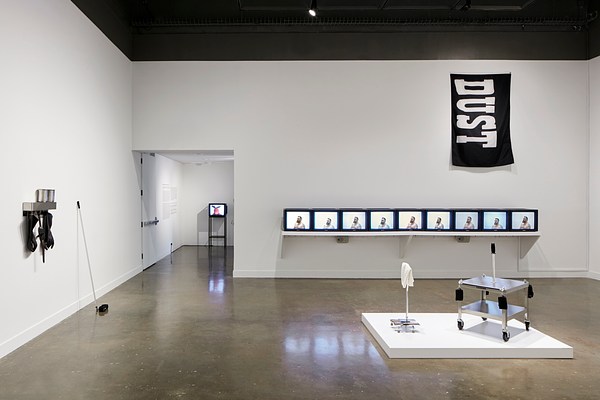 Installation view featuring screens repeating in the center, janitor supplies hanging on the left wall, a metal sculpture in the center, and a black tapestry that says "DUST" on it on the right.