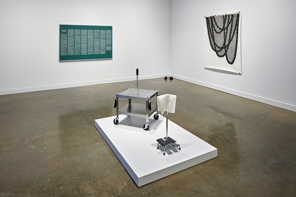 Installation view featuring a metal card on a podium in the center, a green print with white text on the wall, and a tapestry with chain like graphic on the right wall.&nbsp;